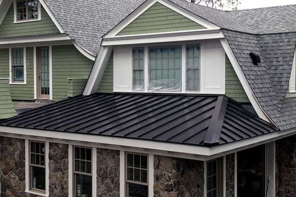 Residential roofing installation featuring a modern black metal roof on a house with green siding and stone accents, showcasing Twin Roofing's quality craftsmanship.