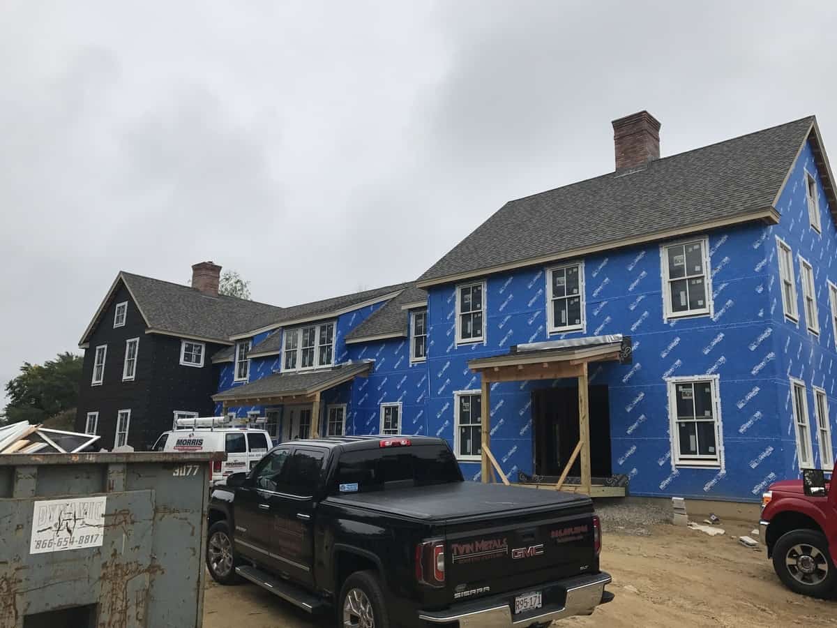 Residential home under construction with blue insulation wrap, showcasing new roofing installation, surrounded by construction vehicles, emphasizing Twin Roofing's commitment to quality and detail in roofing services.